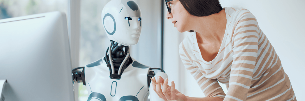 Junge Frau mit schwarzer Brille spricht mit einem Roboter, der vor einem Bildschirm sitzt.