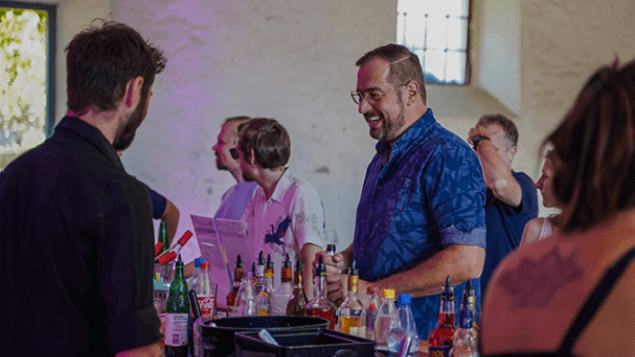 Ein Mann im blauen Hemd steht an der Bar und lacht. Im Hintergrund sind weitere Personen an der Bar zu sehen. Der Barkeeper steht mit dem Rücken zum Betrachter.