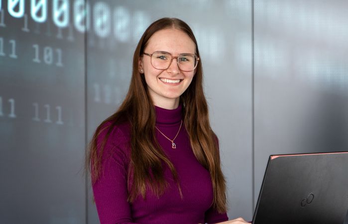 Junge Frau mit lila Pullover arbeitet an einem Laptop und lächelt in die Kamera.