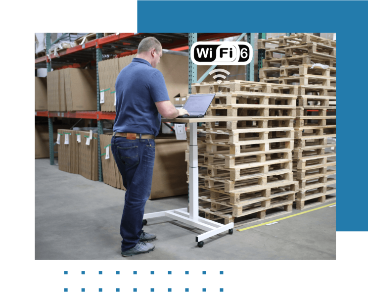 Eine Person arbeitet in einer Lagerhalle an einem Laptop und ist dabei mit WiFi 6 verbunden.