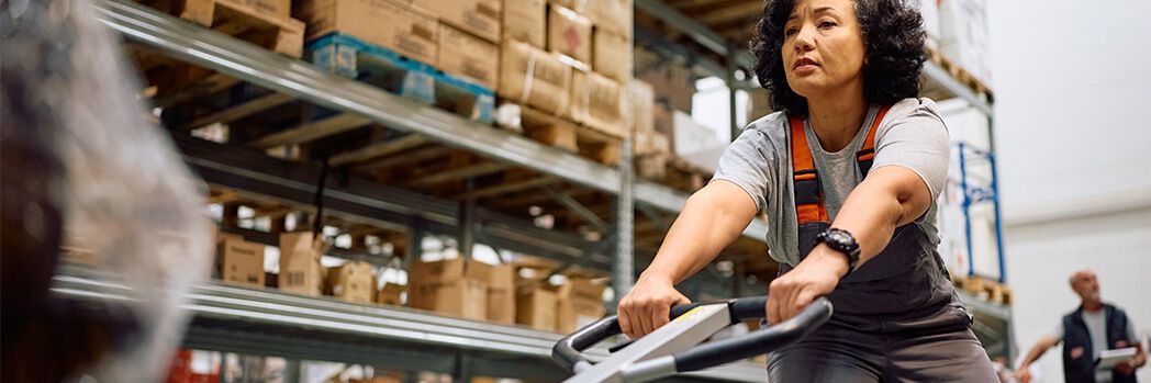 Eine Mitarbeiterin im Lager steuert einen Handhubwagen zwischen Regalen voller Kartons, was die Logistik effizient unterstützt.