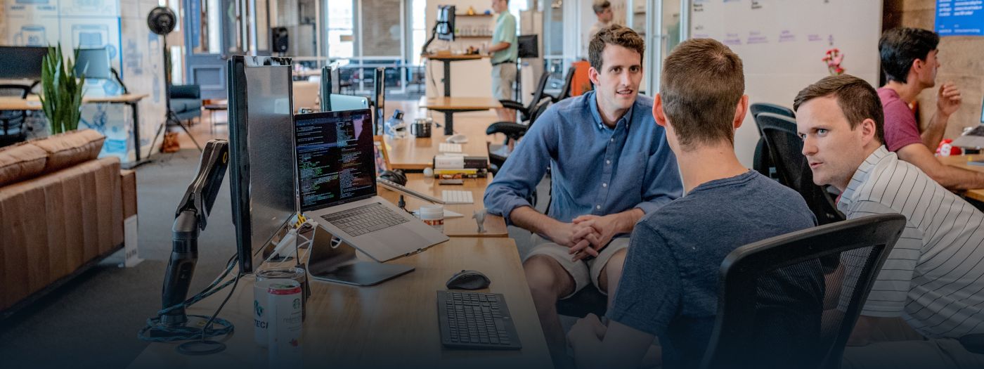 Drei Personen sprechen über ein Projekt während sie vor einem Arbeitsplatz sitzen, auf dem ein Laptop und ein Monitor vorzufinden ist.