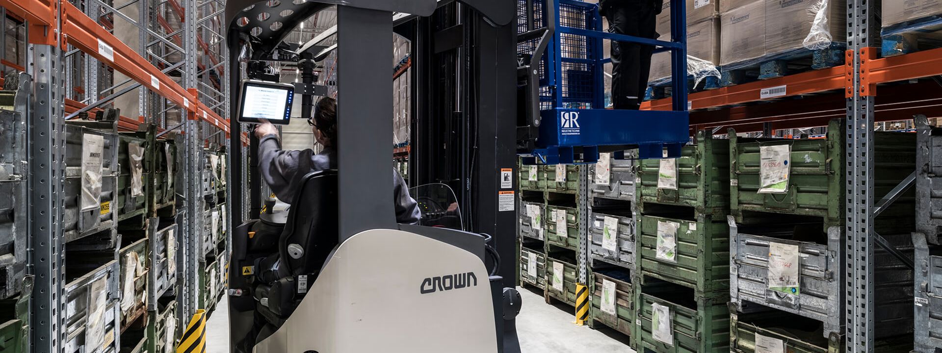 An employee stands on a mobile lifting platform in a warehouse and uses mobile devices.