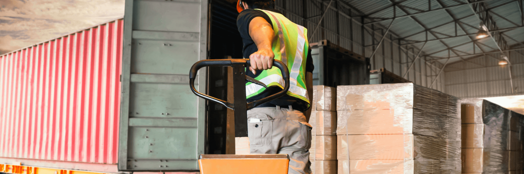 Truck is standing at the ramp with the loading flap open and is being loaded by a worker in a high-visibility waistcoat.