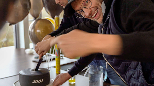 Eine Person schneidet einen Kuchen im Büro an, während zwei Kollegen im Vordergrund stehen.