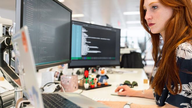 Junge Frau mit roten Haaren arbeitet als Softwareentwicklerin an einem Schreibtisch mit zwei Bildschirmen und einem Laptop vor sich.