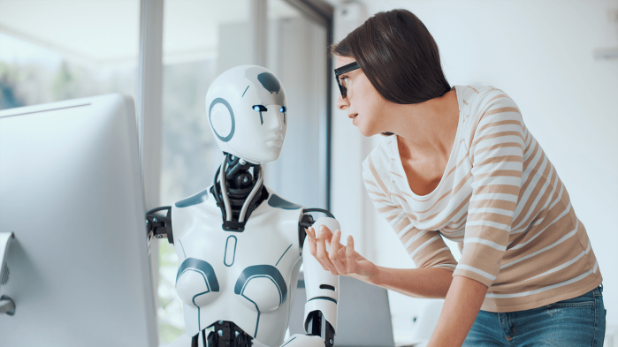 Young woman with black glasses talks to a robot sitting in front of a screen.