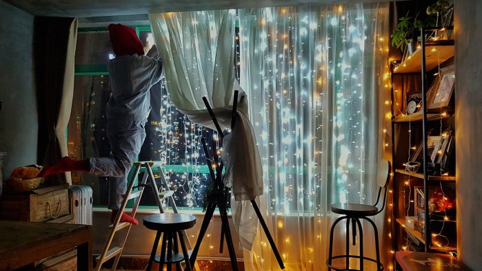 Mann mit Mütze auf dem Kopf hängt Lichterkette an seinem Fenster auf während er auf einer Leiter steht