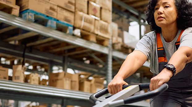 Eine Mitarbeiterin im Lager steuert einen Handhubwagen zwischen Regalen voller Kartons, was die Logistik effizient unterstützt.