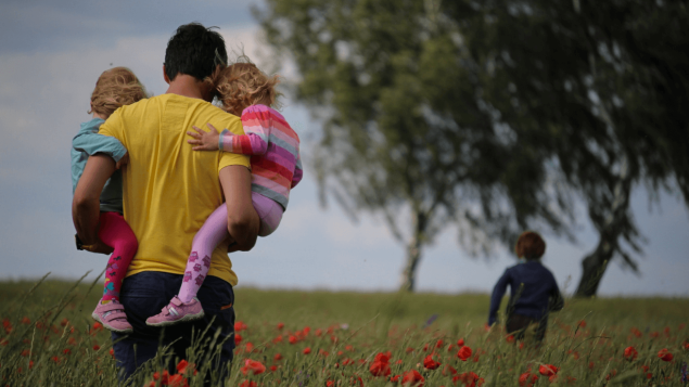 Ein Mann im gelben T-Shirt nutzt die flexiblen Arbeitszeiten und läuft mit seinen drei Kindern über eine Wiese mit Mohnblumen.