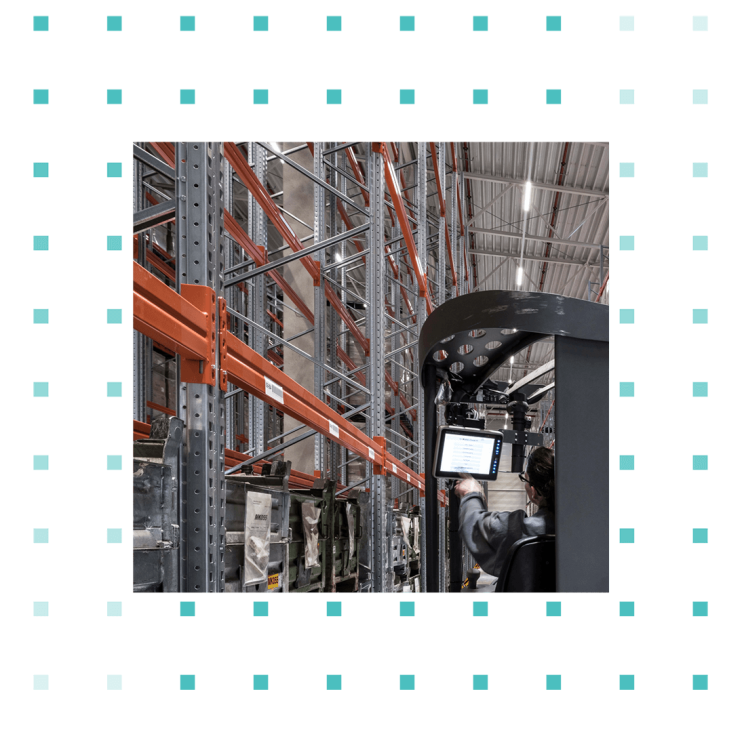 Logistician drives forklift through high-bay warehouse and uses mobile device.