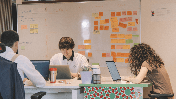 Im Hintergrund ist ein Whiteboard mit bunten Notizen zu sehen, davor sitzen drei Personen an Laptops.