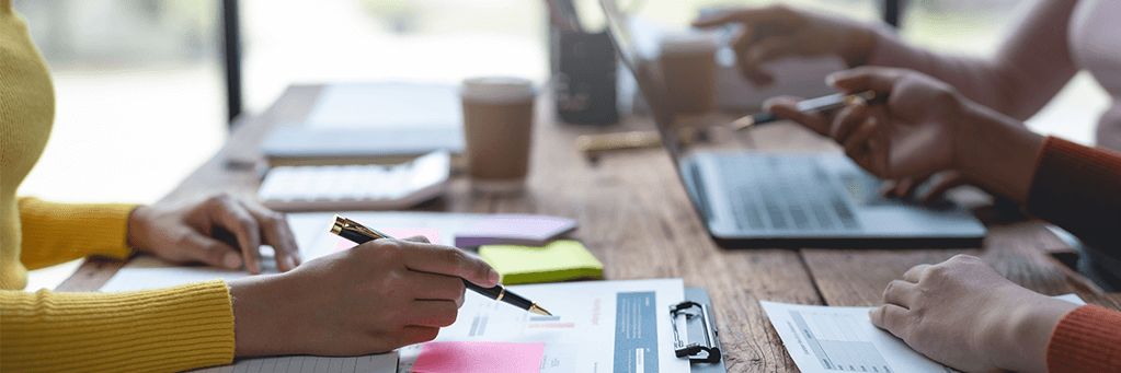 Vier Personen zeigen mit Stiften auf einen Laptop-Bildschirm, während sie gemeinsam an einem Projekt arbeiten.
