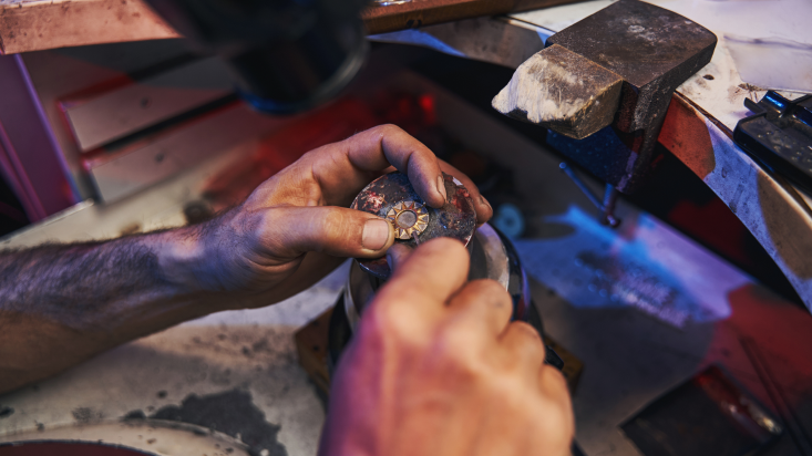 Close-up of hands working on a piece of jewelry at a workstation under a lamp, with tools in the background.