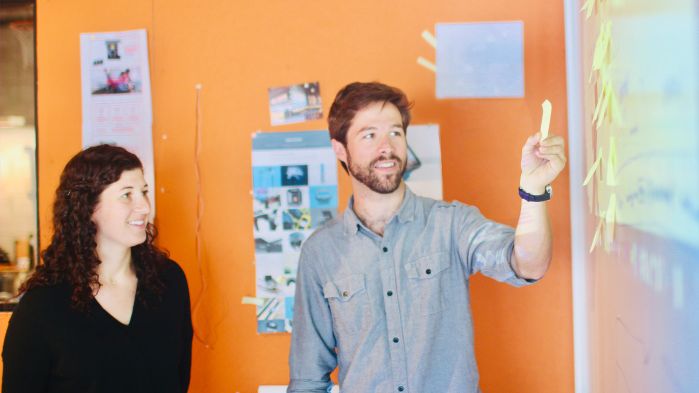 Mann und Frau stehen lächelnd mit Post-it in der Hand an einem Whiteboard