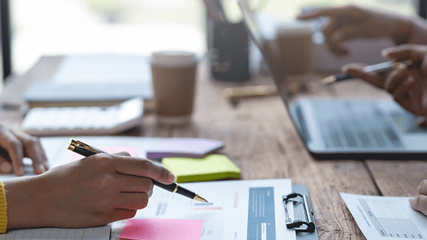 Vier Personen zeigen mit Stiften auf einen Laptop-Bildschirm, während sie gemeinsam an einem Projekt arbeiten.
