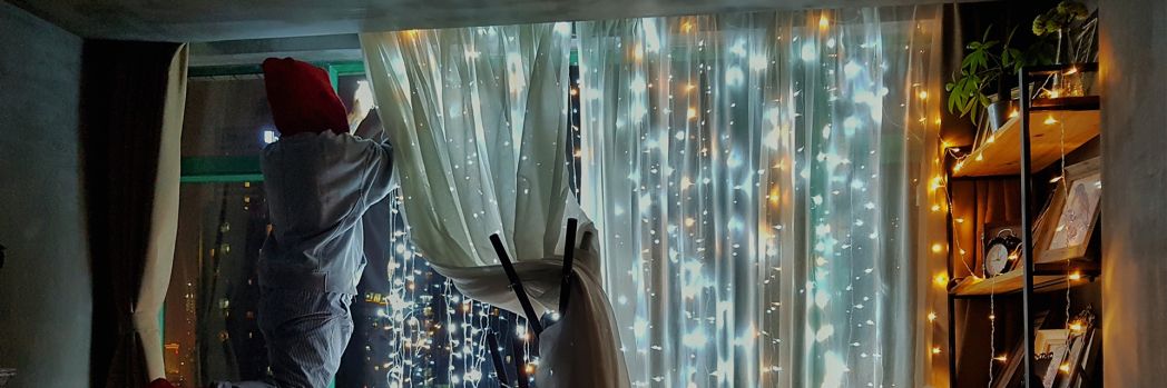 Man with cap on head hangs string of lights on his window while standing on ladder