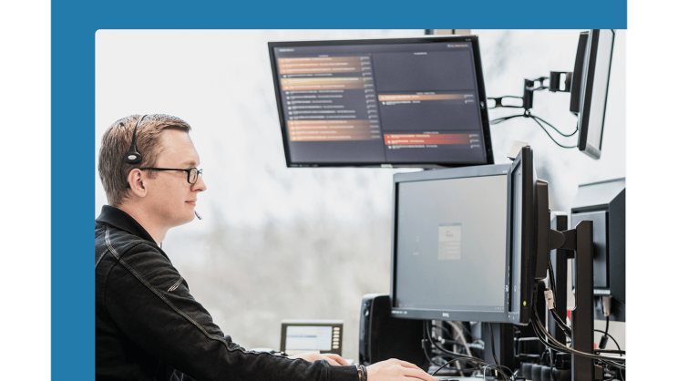 A blond man with a headset and glasses sits at an IT workstation with three screens.