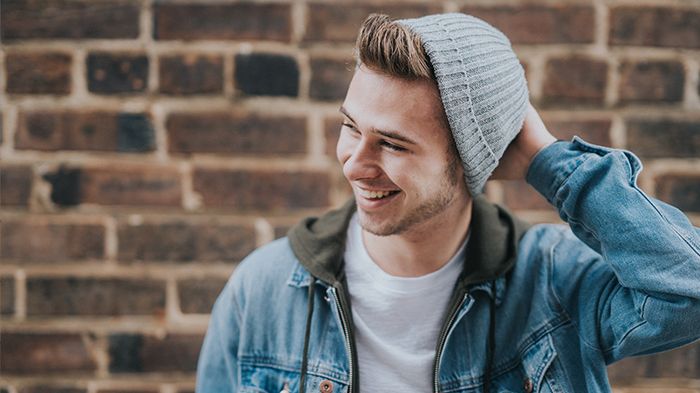 Junger Mann mit Mütze steht vor einer Mauer und lächelt.
