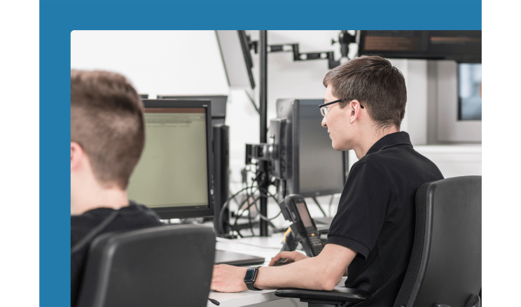Two men in black shirts sit next to each other at a desk with screens on it.
