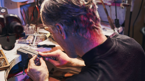 A jeweler in a workshop filing a piece, surrounded by tools and a lamp.