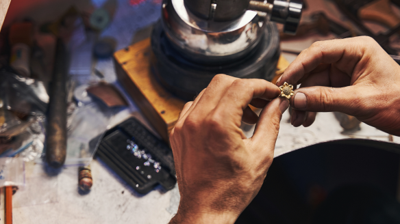 Craftsman's hands working on a small jewelry piece with gemstones, at a well-equipped workstation.