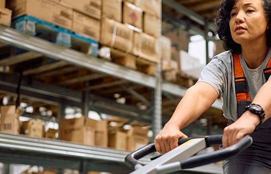 Eine Mitarbeiterin im Lager steuert einen Handhubwagen zwischen Regalen voller Kartons, was die Logistik effizient unterstützt.