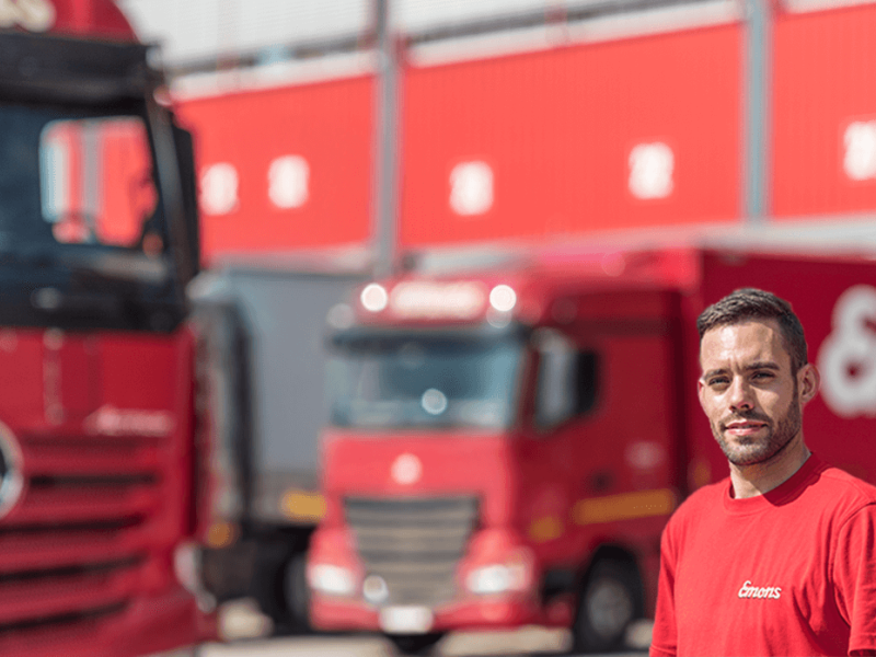 Im Vordergrund ist ein Mitarbeiter der FIrma Emons zu sehen. Im Hintergrund erkennt man zwei rote LKW.