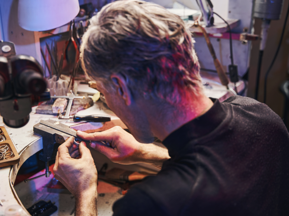 Ein Juwelier in einer Werkstatt, der mit Feilen an einem Werkstück arbeitet, umgeben von Werkzeugen und einer Lampe.