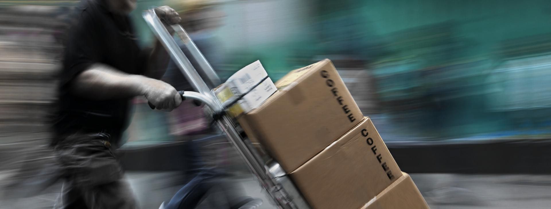 Alles Wichtige zum Transport für Empfänger mit der EIKONA Logistics Cloud. Ohne Fragen, in Echtzeit. Paketbote transportiert drei braune Kartons auf einem Sackkarren.