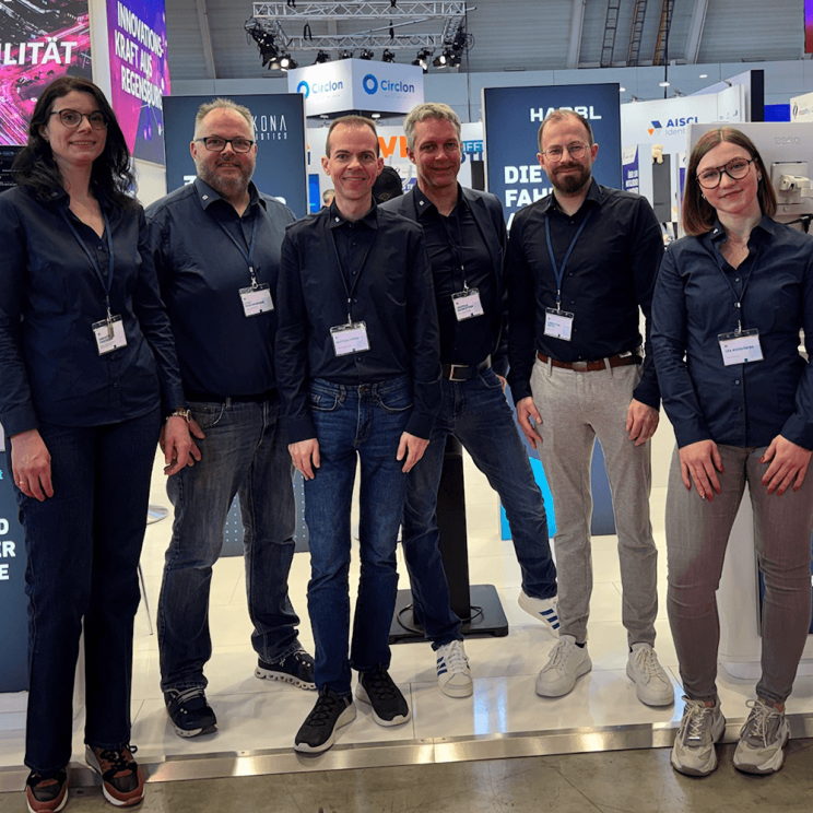 Ein Team von sechs Personen in blauen Hemden lächelt vor einem Messestand und präsentiert ihre Produkte.