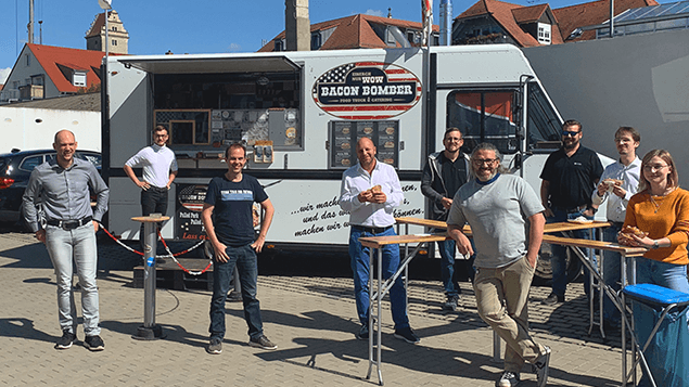Ein Foodtruck steht auf einem Parkplatz, während 9 Personen vor diesem stehen.