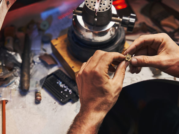 Hände eines Handwerkers, der ein kleines Schmuckstück mit Schmucksteinen bearbeitet, auf einem gut ausgestatteten Arbeitsplatz.