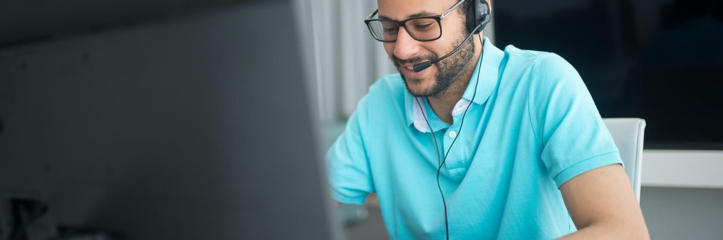 Mann mit türkisem Shirt tippt an seinem Computer während er am Headset telefoniert